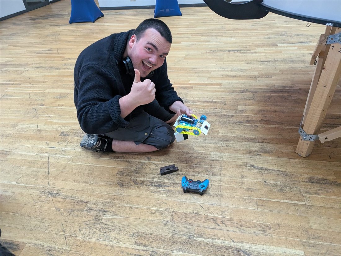 Teilnehmer hält einen selbstgebauten Raupen-Bot, daneben liegt ein Gamepad